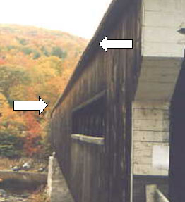 The photo shows a close-up outside long view of a racked, distorted bridge. The white arrows point to the edge of the roof overhang where the bridge bows in or out and is out of square down the longitudinal access.