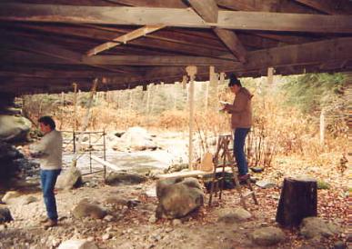 The picture shows two workers underneath the bridge decking taking readings from several dozen dial gauges to measure the response at various locations to known vehicle weights positioned at specific places along the bridge.