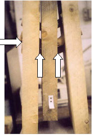 The picture shows a destructive test with three-element specimen to measure direct shear in a three-peg connection. The timber planks were typical spruce and the pegs were 44-millimeter (1.75-inch) diameter rounds. Two white arrows point up vertically, and one points to the right horizontally from the left side showing the trunnel that has failed in bending.