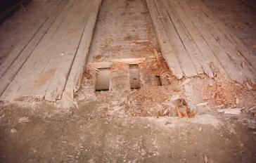 The picture shows a close-up of a bridge deck resulting from poor drainage. The deck planking is rotted away so that the transverse floor beams show. The running planks are also deteriorated and broken near the road surface.