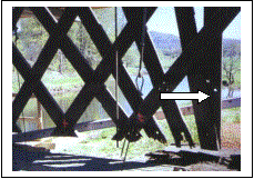 The picture shows a deteriorated lattice truss. The top arrow points to splaying with the empty holes from the original construction while the bottom arrow points to the broken condition of the lattice ends, which was hidden behind steel plates.