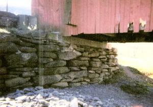 The picture shows a side closeup of the bridge, with deteriorated siding, sitting on a dry-laid stone abutment with a concrete cap.