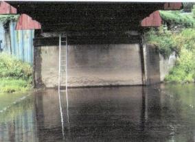 The picture shows the opposite abutment with a ladder ascending the concrete façade in front of the dry-laid stone and steel sheet piling along the wings. The rusty steel frame at the corner of the back wall and transverse across the top of the stem was left over from the 1976 renovation and used for erection, but not for bridge support.