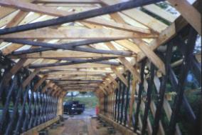 The picture shows the interior of the bridge with the larger beams of the internal bracing and the common rafters that were replaced. The retained darker lateral members contrast with the lighter wood of the replacement members.