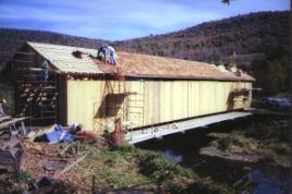 The picture shows a side view of Fitch's bridge being rehabilitated. The decking is extended so staging can be set on it. Staging and access ladders are at the entrance and along the bridge length. Two workers are re-roofing on top of rough-cut nailers and another stands on staging at the entrance.