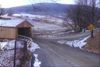 The bridge entrance geometry is sub-standard, yet acceptable to local users. Due to the lack of accident history at the bridge, a heavy timber approach railing system was used. The picture shows two roads converging at the bridge entrance with a wide radius of the approach railing. A chain-link fence shows in the foreground.