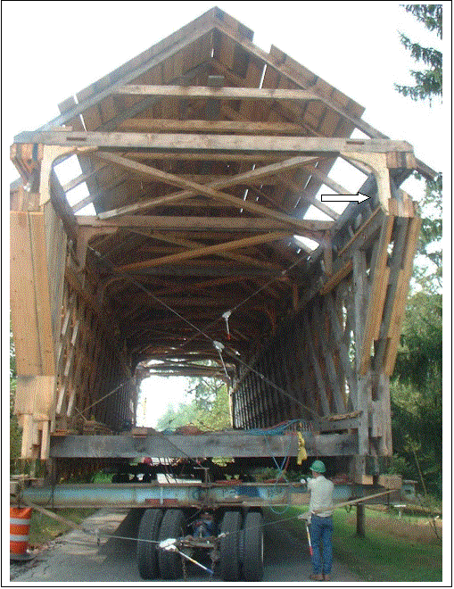 The picture shows a longitudinal view of the replacement Town-lattice truss bridge set on wheels with cross cabling and turnbuckles. The white arrow points to the ship's knees that give good bracing without sacrificing clearance.