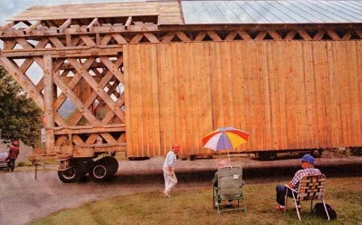 The picture shows a side view of the bridge in transit with the lattice and part of the roofing exposed. Interested observers are sitting in their lawn chairs with umbrellas watching the bridge move past.