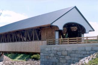 Figure 218 Overall view of bridge showing the overhanging portal, metal roof, outboard sidewalk, main truss, and arch visible though open side, and granite-faced abutment. Photo.