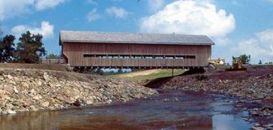 The picture shows a Pratt truss bridge with an extended portal spanning misaligned steep, rocky banks over a bend in the river.