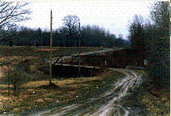 The picture shows a winding road leading to and away from the bridge with the former steel pony-truss bridge poorly aligned.