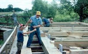 The picture shows four workers assembling the bridge. Two are using a pry bar for the truss assembly.