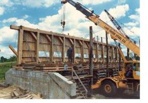 The side view shows the bridge abutment with the north truss being lifted and set in position by a crane. Details reveal the inside superstructure posts and diagonals of the bridge.