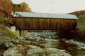 The picture shows the longitudinal view of the long-span bridge (built circa 1883) with natural siding over a river with boulders.