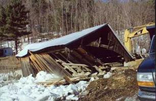 The picture shows the bridge collapsed with snow in the foreground. The roof looks largely intact but the sides have splintered and are leaning outward after thebridge was struck by ice