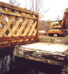 The picture shows a crane in the background dismantling the bridge. The stone abutment with a concrete bearing surface and the lattice truss remain.