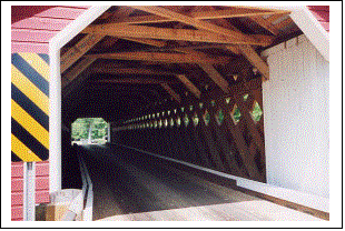 The interior view of the finished bridge shows the Town lattice truss, the reinforced tie beams, the white curbs, and the siding on the portal with a diagonal warning sign.