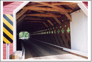 The interior view of the finished bridge shows the Town lattice truss, the reinforced tie beams, the white curbs, and the siding on the portal with a diagonal warning sign.