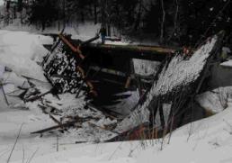The picture shows the demise of the bridge after an excessive snow load. The bridge is tipped on its side with snow surrounding it and the roof is collapsed and broken in multiple pieces.