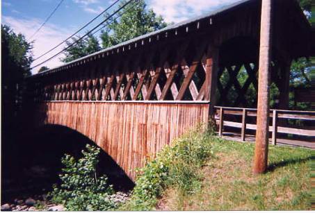 This side view of a Town lattice truss bridge shows plenty of ventilation with the exposed upper lattice, siding that is cut into the shape of an arch and approach rails. It cannot be ascertained if there is really an arch behind the siding, or if it is just an architectural gimmick.