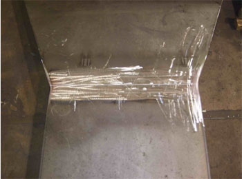 The photo shows a plan view of field specimen FG36K-TF2-TopF-FCM with the weld oriented horizontally. The specimen is comprised of a wider plate to the top and a more narrow plate to the bottom of the photo. The centerlines of the plates are aligned.