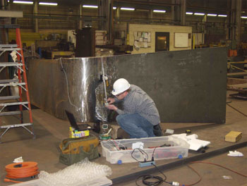 The photo shows a plan view of field specimen FG40M-TF1-Curved-FCM with the weld oriented vertically. The specimen is comprised of curved plates of unequal thickness and equal width. The thicker plate is to the right in the photo. A man is crouched in front of the specimen. He has the P-scan system mounted on the specimen and is managing the ultrasonic transcducer with both hands. He is looking at the lap top computer positioned on a box in front of him. A large plastic box is open beside him with cords coming out of it to power strips and the computer. He is wearing jeans, a plaid shirt and a white hardhat.