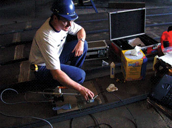 The picture shows typical scanning activities during the P-scan system field testing at High Steel Structures, Inc. A man wearing a white T-shirt, jeans, and a blue hardhat is crouched next to the P-scan system. His hand is on the ultrasonic transducer. He is looking in the direction of the laptop computer which is partially visible opposite of him. Equipment cases and boxes are open on the floor around him.