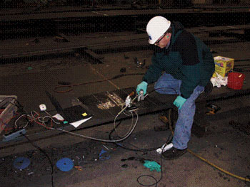 The picture shows typical scanning activities during the P-scan system field testing at Stupp Bridge Company. A man is positioned partially over what appears to be a long panel of steel. The P-scan system is positioned on a welded piece. The man has the ultrasonic transducer in his right hand. He is wearing a green and black coat, green gloves, jeans, and a white hardhat.