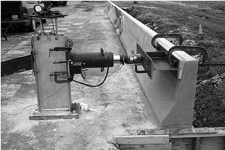 Figure 132. Photo. Test setup for static load tests on safety-shaped barriers. This figure is a full view of the safety-shaped barrier. It is connected to the deck. On top of the deck sits the the hydraulic ram. A piston runs from this device to a steel I-beam attached to timber. The steel plate of the I-beam and timber are clamped to the top right edge of the parapet.