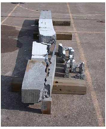 Figure 72. Photo. Damage dominates one side of the beam impacted at 15.9 kilometers per hour (9.9 miles per hour). This is a post-test photo of the broken beam. It is broken into three main pieces, two on one side of the beam where extensive debonding is evident. The pieces are sitting on wood blocks resting on the ground.