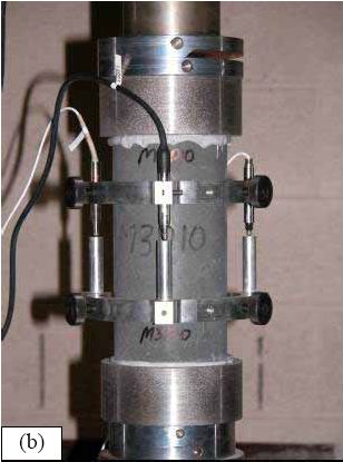 Photos. (a) Notched cylinder and (b) testing of an unnotched cylinder. (a) shows a 102-millimeter diameter cylinder after it has had a notch milled into its circumference at midheight. (b) shows an unnotched cylinder undergoing direct tension testing. This cylinder has been epoxied into the testing machine and an axial displacement measuring device has been attached to the specimen.