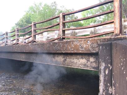 Figure 2. Photo. Bridge fascia immediately after fire. This photo shows an elevation view of the bridge fascia immediately after the fire has been extinguished.  Surface spalling is apparent on the vertical surface of the exterior beam, on the vertical surface of the curb, and on the face of the abutment. The railing on the bridge has also been damaged, with the horizontal rails being bowed by the intense heat of the fire.