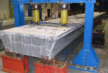 This photo illustrates the setup used in the shear testing of the ultra-high performance pi-girder. Two hydraulic jacks are applying a load to the top surface of the girder, and each end of the girder is supported by rollers. One end of the girder is seen in the foreground, and the bottom of the bulbs is supported by rollers sitting on fixed abutments. The angle of the photo shows both the end of the pi-girder as well as part of the side and top of the girder.