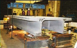 This photo illustrates the test setup used in the transverse flexure testing of the ultra-high performance concrete pi-girder. Two hydraulic jacks are applying a load to the top surface of the girder near midspan, and each end of the girder is supported by elastomeric pads resting on fixed abutments. The oblique angle from which the photo was captured allows for viewing of both the end of the pi-girder as well as a side face.
