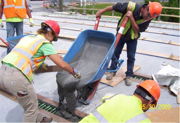 Figure 1. Photo. Placement of UHPC into longitudinal connection between deck-bulb-tee girders. (Photo courtesy of New York State Department of Transportation.). This photograph shows workers casting fluid ultra-high performance concrete (UHPC) from a wheelbarrow into the void space between the top flanges of two deck-bulb-tee prestressed concrete girders. The rebar extends from the girders into the void. The UHPC is self-consolidating.