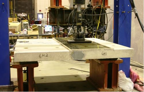 Figure 3. Photo. Field-cast UHPC longitudinal connection cyclic test setup. This photograph shows the elevation view of the test setup used for the cyclic testing of the longitudinal connection specimens. Roller supports are to the left and right, the test specimen is in the center of the photo, and the cyclic loading actuator is above the specimen near midspan. The connection is in the middle of the span. A pond of water is on top of the specimen to allow for assessment of connection leakage during the cyclic loading.