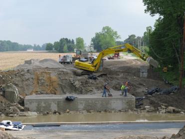 Photo showing the construction of a geosynthetic reinforced soil (GRS) abutment and wing walls. A core of native soil is shown behind the compacted reinforced fill (forming the cut slope) upon which a track hoe sits and operates.