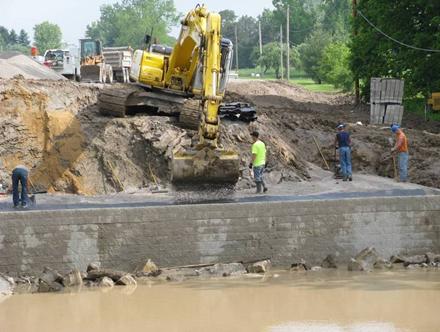 Photo showing a typical labor crew of 4 workers with a trackhoe centrally located on top of the cut slope. The trackhoe is dump spreading a layer of gravel on the geotextile. The workers are compacting the gravel. The abutment is being built in slightly flooded water condition with the water level to the top of the riprap.