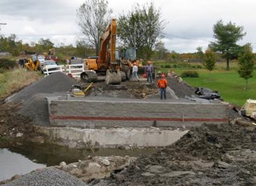 Photo showing the excavator positioned inside the wall area for easy placement of fill, block, and other materials from the staging area to the geosynthetic reinforced soil (GRS) abutment. The GRS abutment is being built behind an existing concrete abutment wall. The top of the existing concrete was removed to allow for the added width of the new bridge.