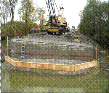 Photo showing a face view of a geosynthetic reinforced soil (GRS) abutment with a superelevation that is built behind an existing concrete abutment wall. The top of the concrete wall was removed and capped with concrete. Formwork is shown for this concrete cap. A crane is onsite behind the abutment to place the beams.