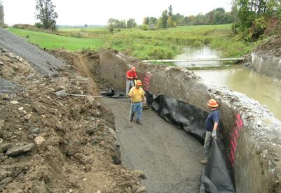Photo showing the construction of the reinforced soil foundation (RSF) behind the existing concrete abutment. There are three laborers compacting, holding, and raking out the fill. The concrete wall has spray painted lines at 8-inch increments as a guide for the compacted layers of fill.