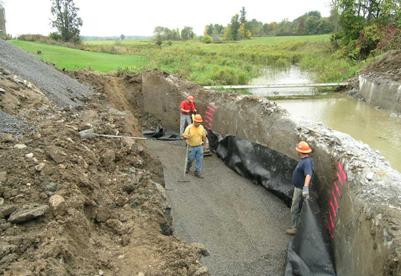 Photo showing the construction of the reinforced soil foundation (RSF) behind the existing concrete abutment. There are three laborers compacting, holding, and raking out the fill. The concrete wall has spray painted lines at 8-inch increments as a guide for the compacted layers of fill.