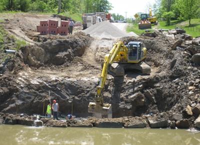 Photo showing the construction of a reinforced soil foundation (RSF). The excavator is positioned on top of the stable cut slope above the abutment. The RSF excavation is below the river level. The workers are in the pit, building the RSF. One worker is measuring elevation with a survey level rod. The abutment is being built in slightly flooded water conditions with the water level to the top of the riprap.