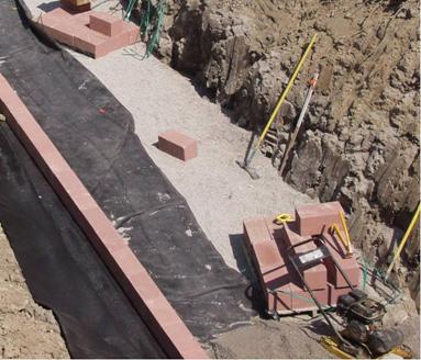 Photo showing the placement of the first course of solid concrete modular block directly on the geotextile-encapsulated reinforced soil foundation (RSF). The solid blocks are red and will be embedded within the riprap. There is an assortment of tools to spread the gravel and a vibratory plate compactor shown in the photo.