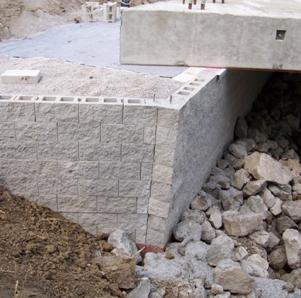 The photo shows a modular block geosynthetic reinforced soil (GRS) abutment built with a obtuse angle wing wall (greater than 90 degrees). The use of rectangular modular blocks makes it necessary to cut the blocks at angle, creating a vertical seam at the corner where the wing wall and abutment wall meet. The photo also illustrates the placement of the riprap at the abutment face and placement of the concrete box beams directly on the GRS abutment.