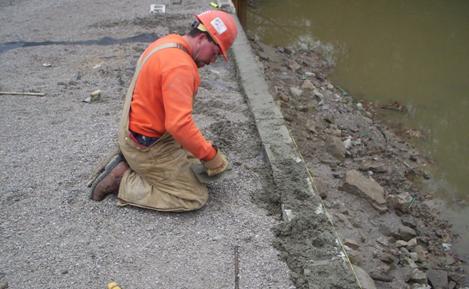 The photo shows a laborer filling the top three courses of a modular facing block with concrete. In the bottom of the photo is a 0.5 inch rebar dowel, which is also placed in the hollow-core block as part of the grouting procedure.
