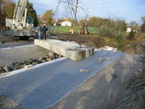 This photo illustrates placement of the concrete box on the geosynthetic reinforced soil (GRS) abutment bearing seat. The photo is from afar, with two men standing on the box beam stretched across a gap.