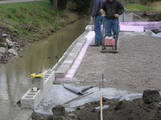 The photo  shows a row of 4-inch foam beneath a row of solid 4-inch concrete blocks directly behind the facing block. Workers are compacting the geosynthetic reinforced soil (GRS) backfill to prepare the area to construct the beam seat.