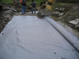 The photo shows construction of the first 4-inch wrapped layer in the beam seat. A track hoe is spreading a 4-inch-thick layer of gravel from the face of the wall backwards. Laborers are smoothing the gravel layer level 