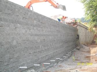 Photo showing the construction of a geosynthetic reinforced soil (GRS) wall to repair a panel-faced mechanically stabilized earth (MSE) wall. The repair GRS wall is built with concrete masonry unit (CMU) facing blocks. The base of the GRS wall has a row of plastic pipes to facilitate drainage. The top of the picture shows several laborers assembling the wall with the aid of a track hoe.